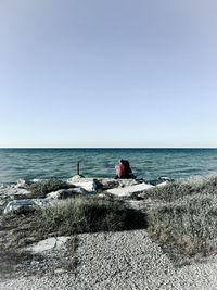 People sitting on rock looking at sea against clear sky