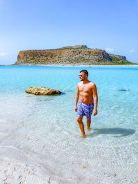 Full length of shirtless man on rock at beach against blue sky