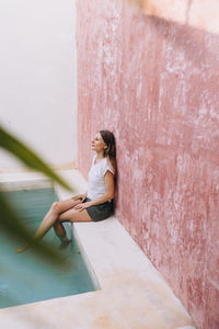 Blond, caucasian woman leaning against a colored wall by a pool, high angle view