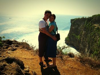 Portrait of couple embracing on cliff by sea against sky