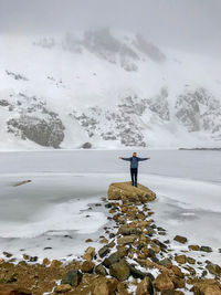 Man with arms outstretched standing on rock against lake during winter