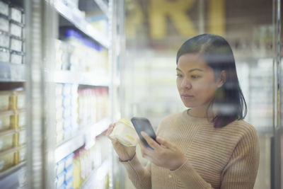 Woman in supermarket standing in front of fridge and using cell phone