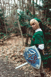 Side view of young man in elf costume with sword and shield standing at forest