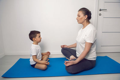 Grandmother and her grandson are sitting on a yoga mat in a white apartment at home and meditating 