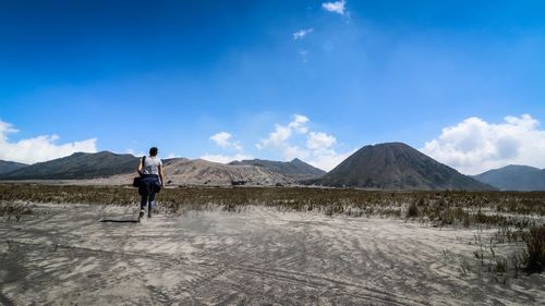 Rear view of woman walking on field by mountains against blue sky