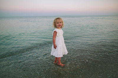 Curly-haired girl standing barefoot by the calm sea at sunset