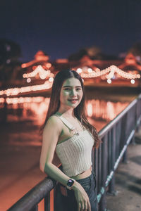 Portrait of young woman standing by railing at night