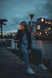 Portrait of young woman sitting on chair at riverbank in city during dusk