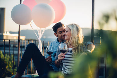 Couple sitting with wine by balloons against sky