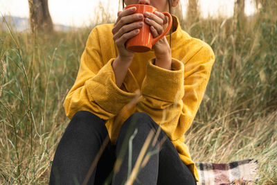 Cropped unrecognizable tranquil female sitting in field in autumn and drinking hot beverage while enjoying weekend