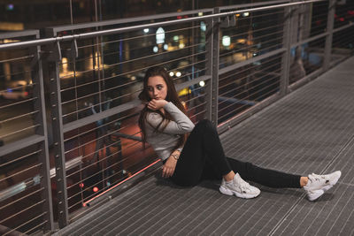 Full length of woman looking away while sitting on footbridge in city at night