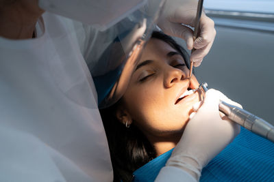 Crop professional dentist in uniform with medical mask drilling tooth of calm woman with help of assistant