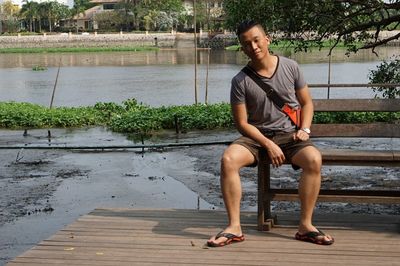 Full length portrait of man sitting on bench against river 