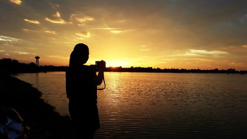 Silhouette man photographing by lake against sky during sunset