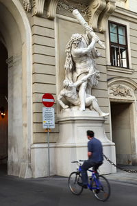 Rear view of man riding bicycle on road against sculpture at building