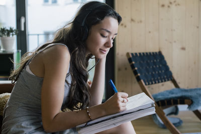 Young woman listening to music and writing in book while sitting at home