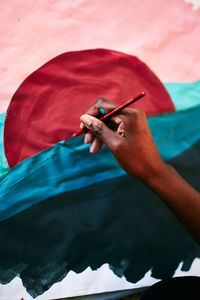 From above of crop anonymous black female with paintbrush drawing colorful picture on paper while sitting on floor at home