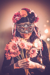 Young man wearing masquerade mask while holding flowers against wall
