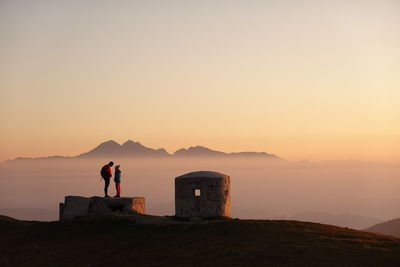 Father and daughter standing on rock against mountain and sky during sunset