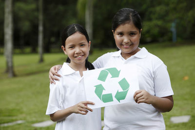 Portrait of smiling friends holding recycling symbol while standing on field