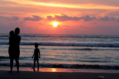 Silhouette mother with her children standing on shore at beach against sky during sunset