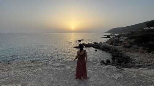 Rear view of woman standing at beach against sky during sunset