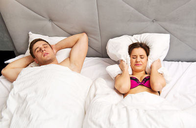 Young man sleeping while woman covering ears with pillow on bed at home