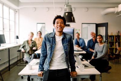 Portrait of young businessman leaning on table while coworkers sitting in creative office