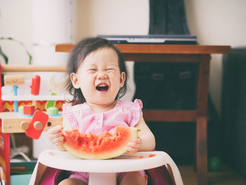 Baby girl holding watermelon while sitting on high chair at home