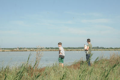 Father and son standing on grassy field by lake against sky