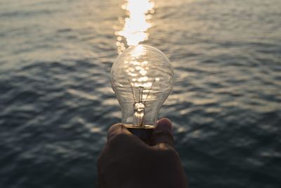 Cropped hand holding light bulb against sea with reflection