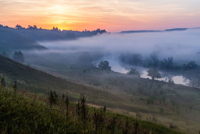 Summer morning fog at the river behind a wild green slope and far forest