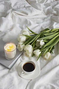 Cozy still life composition with coffee, candle and white tulips on bed.