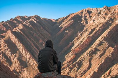 Rear view of man sitting on rocky mountains against clear sky