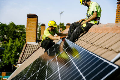 Engineer sitting on roof with colleague installing solar panel