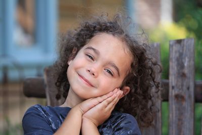 Close-up portrait of smiling girl leaning face on clasped hands outdoors