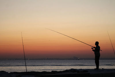 Man fishing in sea against sky during sunset