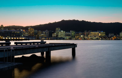Illuminated buildings by river against sky at sunset
