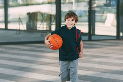 Portrait of smiling boy holding basketball ball while walking on road