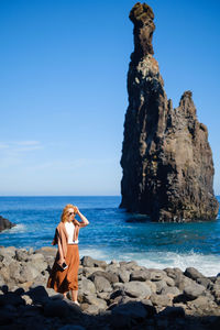 Woman wearing sunglasses standing on beach against rock formation and sky