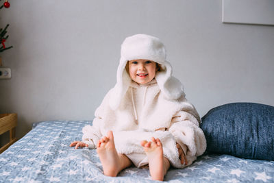 Cute child y on the bed in cozy adult clothes and a hat with earflaps.