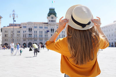 Back view of pretty girl holding hat in trieste, italy. beautiful young woman visiting europe.