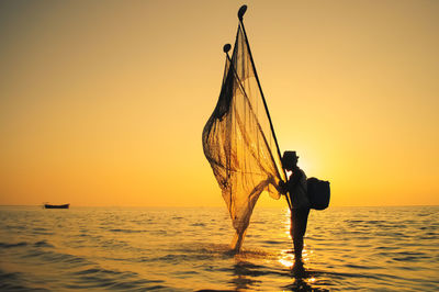 Fisherman with net standing in sea against orange sky during sunset