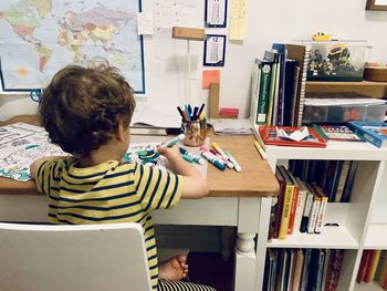 Rear view of boy sitting on table in kids room and drawing on paper