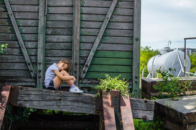 Sad girl sitting on abandoned wooden cargo container against sky