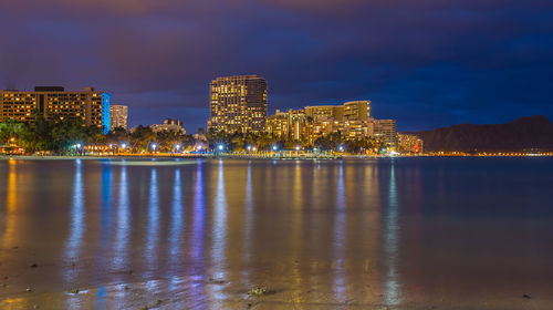 Illuminated buildings by river against sky at night