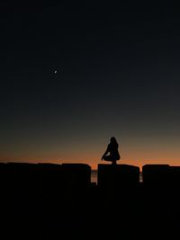 Silhouette woman crouching on retaining wall against clear sky during sunset