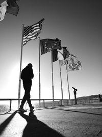 Silhouette man walking on bridge by various flags against sky during sunny day