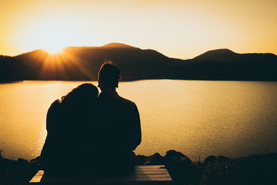 Rear view of silhouette couple sitting by lake against sunset sky