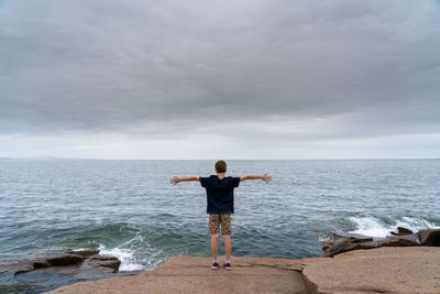 Rear view of carefree boy with arms outstretched standing by sea against cloudy sky